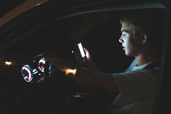 A teenage driver looking at a phone while driving at night, highlighting the dangers of distracted driving among young motorists.