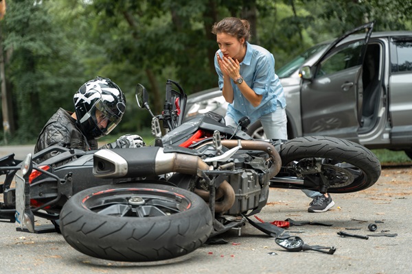 Motorcyclist sitting on the ground next to a fallen motorcycle after a crash, with a concerned woman nearby and a damaged gray vehicle with an open door in the background.
