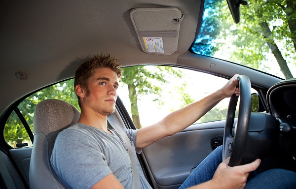 Teen boy sitting in the driver’s seat of a car with one hand on the steering wheel, looking ahead while driving.