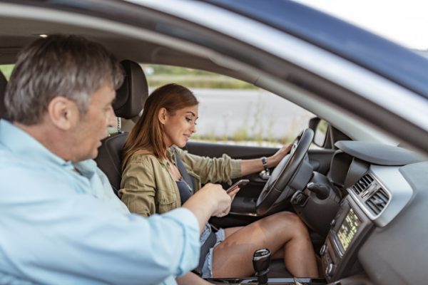 A young teen driver looks at her phone while a man in the passenger seat points at it with disapproval to indicate she should not be on the phone while driving.