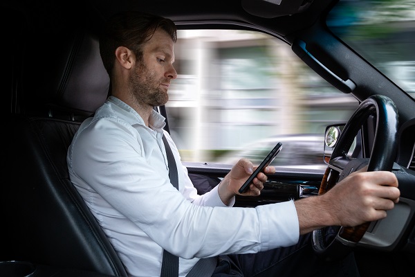 A middle aged man looking down at a smartphone, texting while driving with motion blur outside the car's windows.